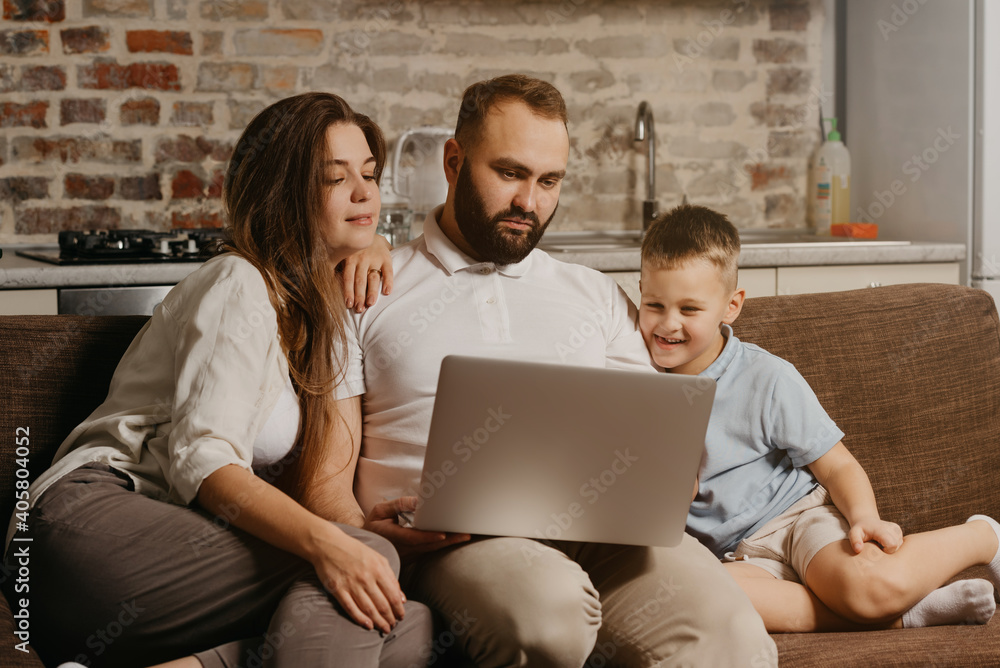 A father with a beard is working remotely on a laptop while his happy son and wife are staring at the screen. Dad is working online on a computer between relatives on the sofa at home in the evening A father with a beard is working remotely on a laptop while his happy son and wife are staring at the screen. Dad is working online on a computer between relatives on the sofa at home in the evening