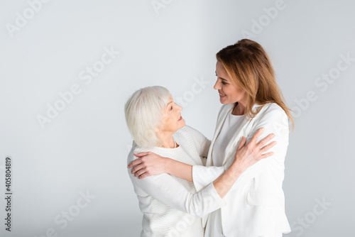 senior mother and happy daughter looking at each other while hugging isolated on grey