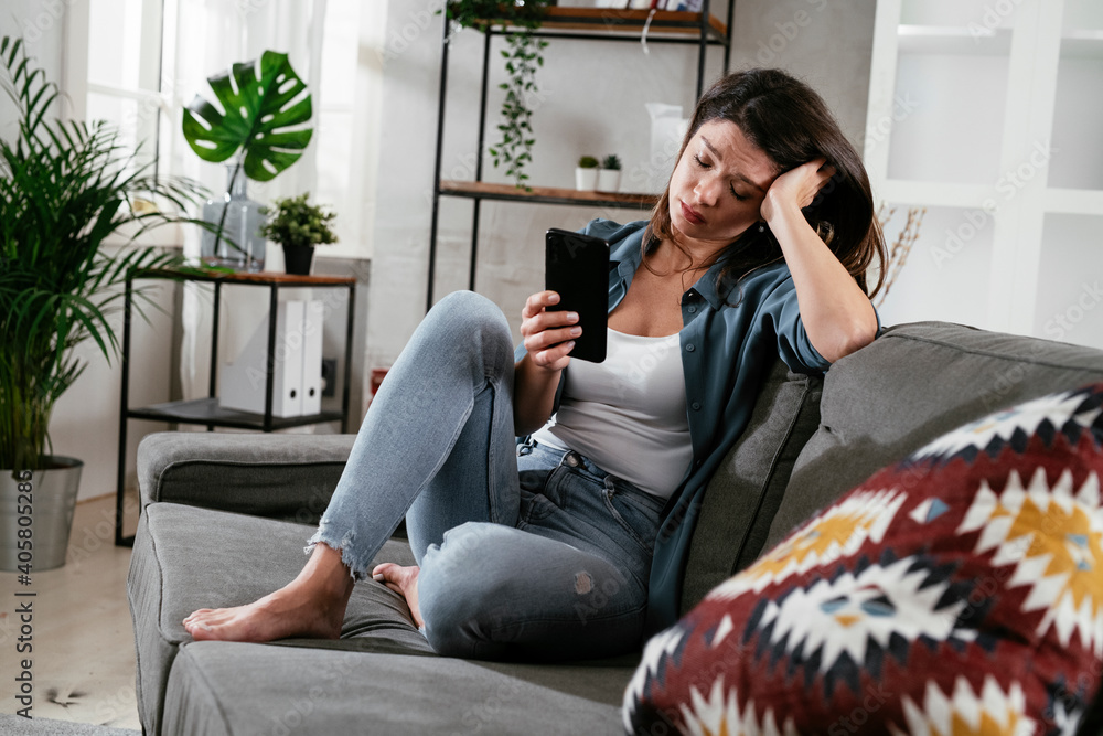 Obraz premium Sad woman sitting on the couch, using the phone. Upset woman waiting for a phone call...
