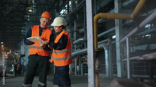 Two maintenance engineers man, woman inspect. Work heavy industry manufacturing factory. Mechanic, female engineer talking in plant. Young employees wearing helmets are working industrial factories