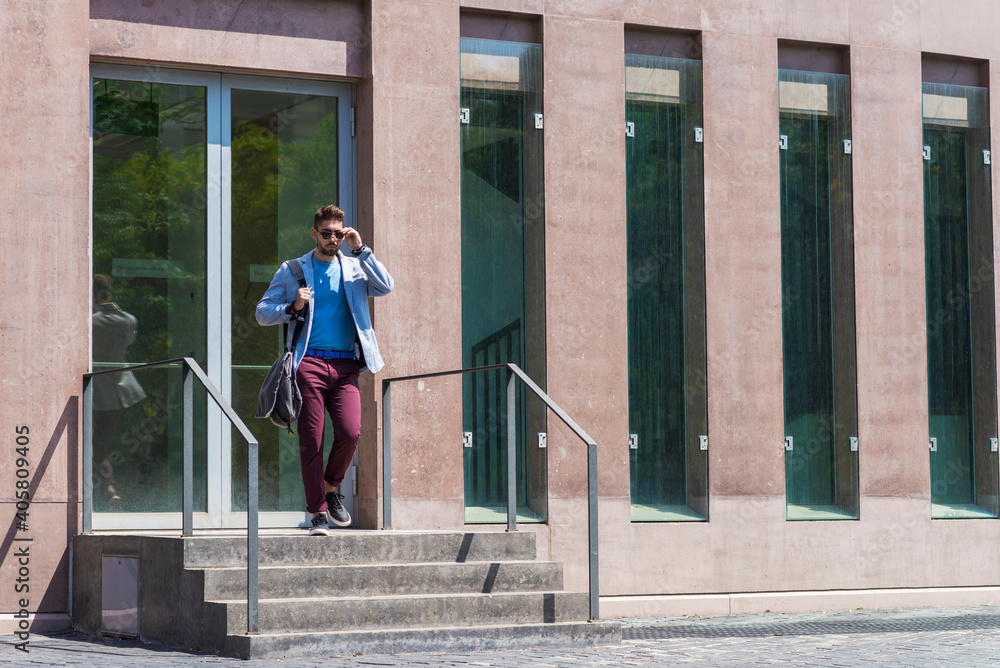 Young businessman walking next to office buildings while holding a shoulder bag outdoors Young businessman walking next to office buildings while holding a shoulder bag outdoors