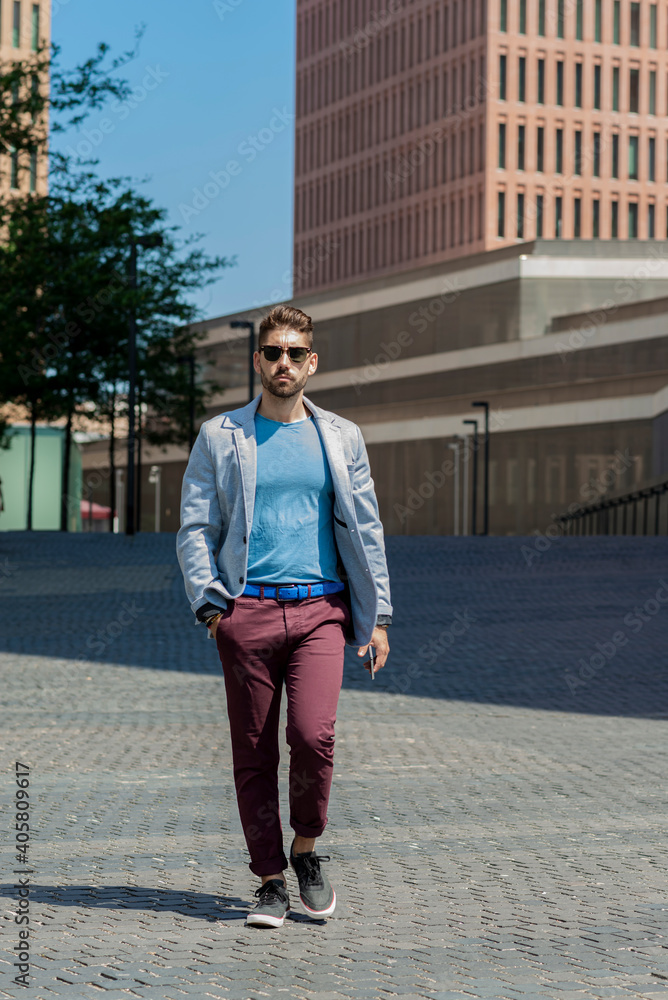 Portrait of a young bearded man with sunglasses, model of fashion, in urban background wearing casual clothes while walking with hand on pocket
