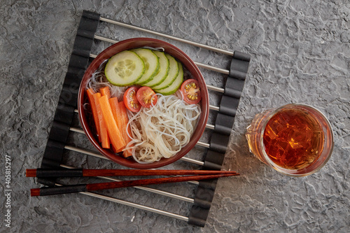 Asian noodle soup, Ramen with vegetables in a bowl on a stand for hot dishes. Gray textured background top view. Ingredients carrots, cucumbers, tomatoes, noodles.