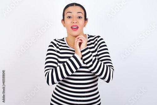 Positive Young beautiful woman wearing stripped t-shirt against white background smiles happily, glad to receive pleasant news from interlocutor, keeps hands together. People emotions concept.