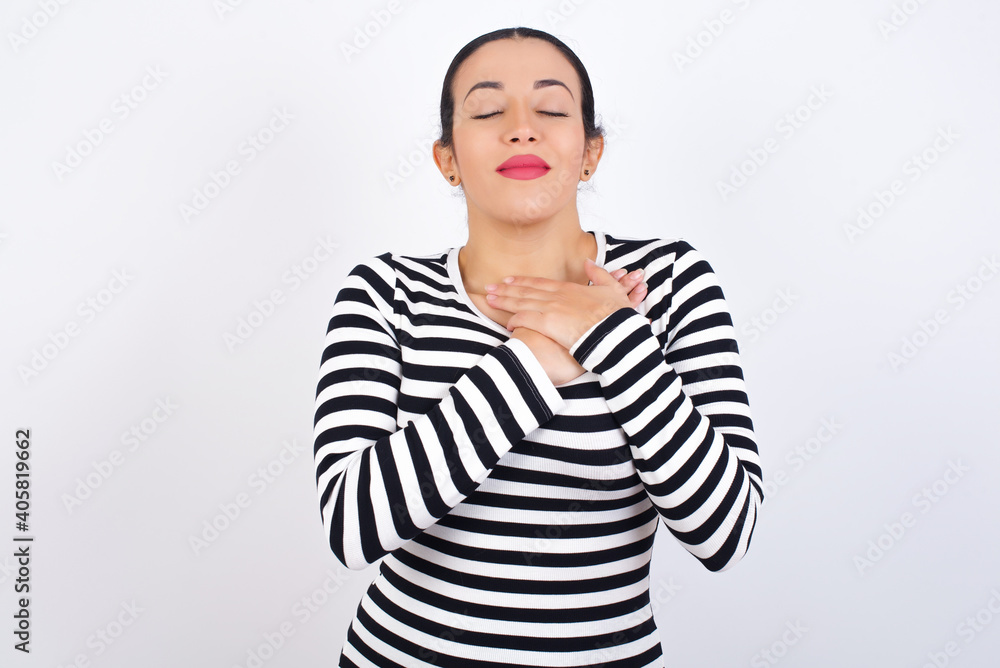 Young beautiful woman wearing stripped t-shirt against white background smiling with hands on chest with closed eyes and grateful gesture on face. Health concept. Young beautiful woman wearing stripped t-shirt against white background smiling with hands on chest with closed eyes and grateful gesture on face. Health concept.