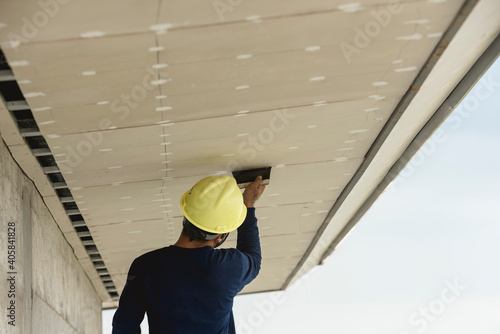 A man applies putty to gypsum roof overhang of a commercial building. Wearing a yellow safety helmet.