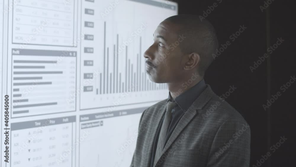 Pensive black professional investor analyzing trading statistics, thinking financial charts. Businessman looking at graphs on big presentation screen, using gadget. Investment concept Pensive black professional investor analyzing trading statistics, thinking financial charts. Businessman looking at graphs on big presentation screen, using gadget. Investment concept