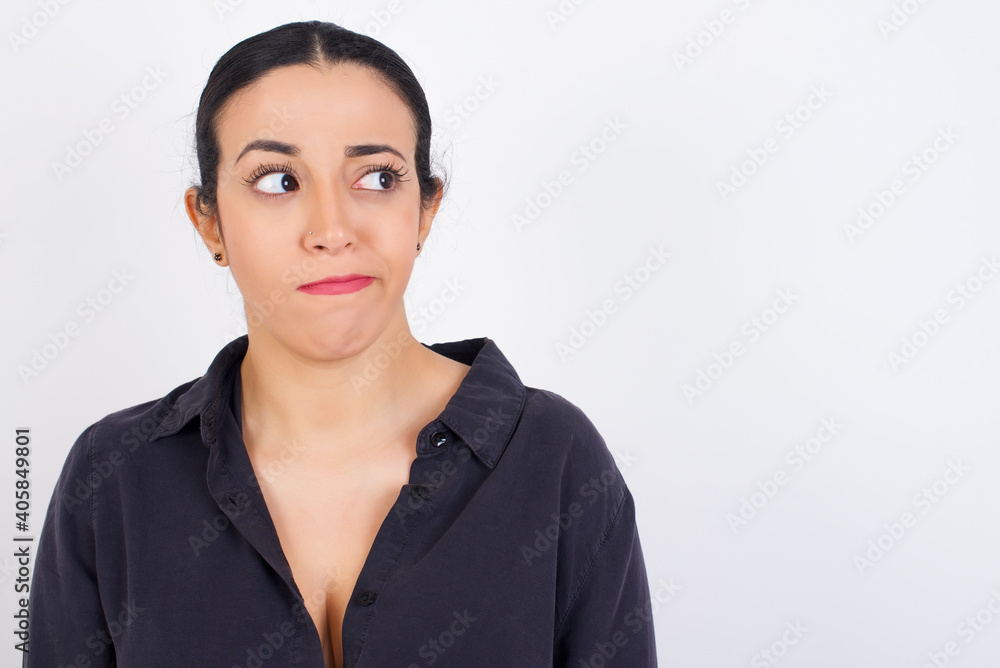 Amazed puzzled young beautiful Arab woman wearing gray dress against white studio wall, curves lips and has worried look, sees something awful in front. Amazed puzzled young beautiful Arab woman wearing gray dress against white studio wall, curves lips and has worried look, sees something awful in front.