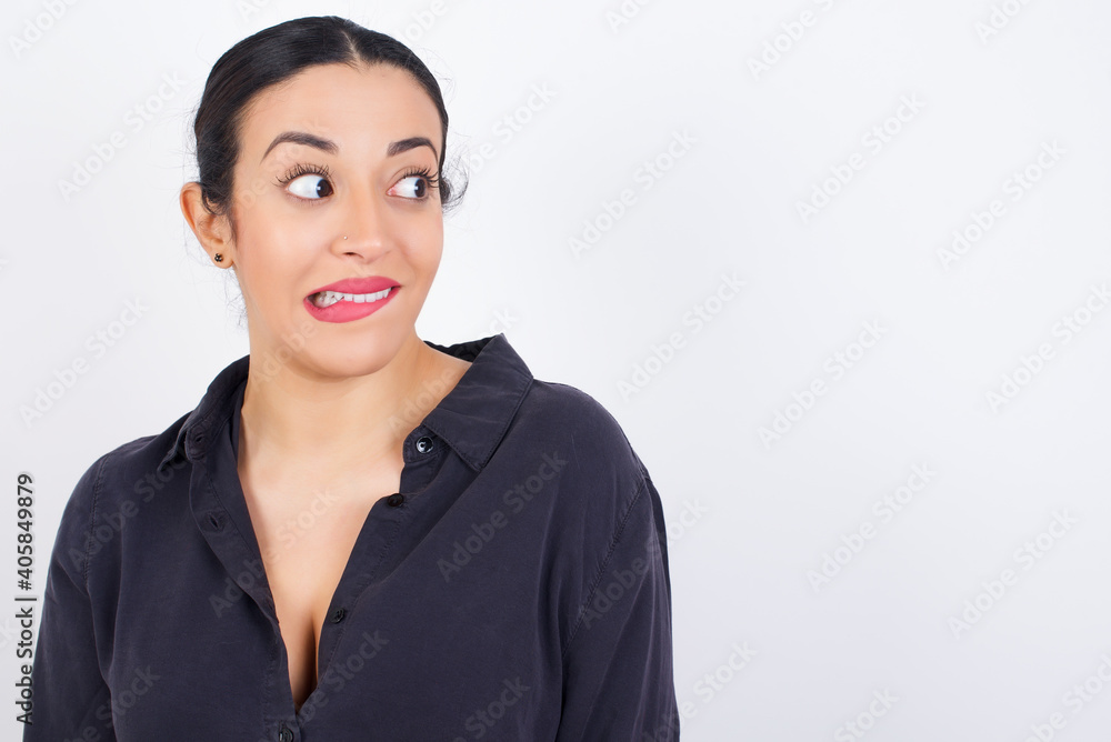 young beautiful Arab woman wearing gray dress against white studio wall with thoughtful expression, looks away keeps hands down bitting his lip thinks about something pleasant. young beautiful Arab woman wearing gray dress against white studio wall with thoughtful expression, looks away keeps hands down bitting his lip thinks about something pleasant.