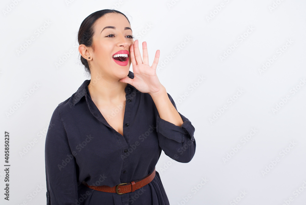 young beautiful Arab woman wearing gray dress against white studio wall profile view, looking happy and excited, shouting and calling to copy space. young beautiful Arab woman wearing gray dress against white studio wall profile view, looking happy and excited, shouting and calling to copy space.