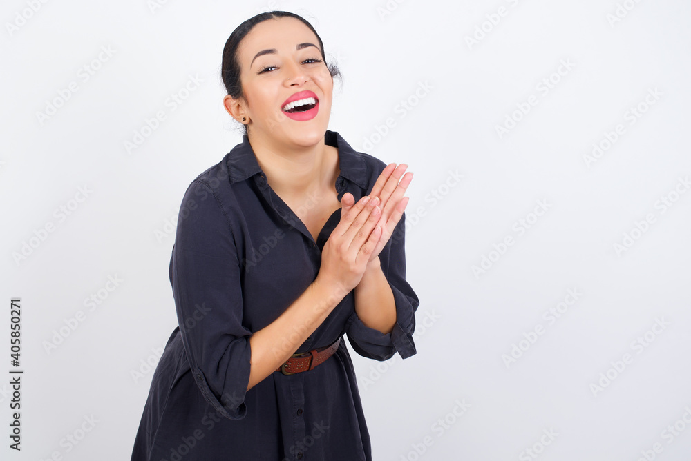 young beautiful Arab woman wearing gray dress against white studio wall, feeling happy, smiling and clapping hands, saying congratulations with an applause. young beautiful Arab woman wearing gray dress against white studio wall, feeling happy, smiling and clapping hands, saying congratulations with an applause.
