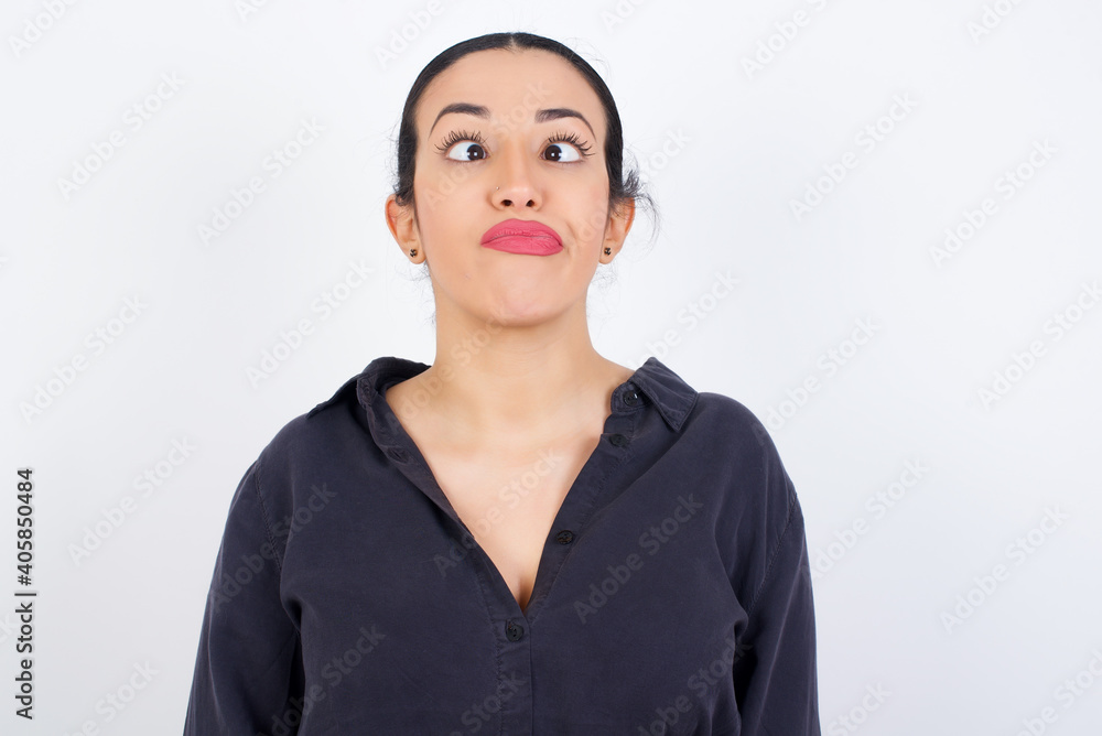 young beautiful Arab woman wearing gray dress against white studio background crosses eyes, puts lips, makes grimace with awkward expression has fun alone, plays fool. young beautiful Arab woman wearing gray dress against white studio background crosses eyes, puts lips, makes grimace with awkward expression has fun alone, plays fool.
