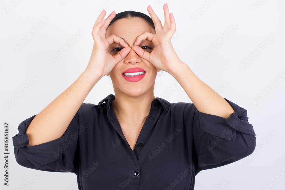 Playful excited young beautiful Arab woman wearing gray dress against white studio wall showing Ok sign with both hands on eyes, pretending to wear spectacles. Playful excited young beautiful Arab woman wearing gray dress against white studio wall showing Ok sign with both hands on eyes, pretending to wear spectacles.