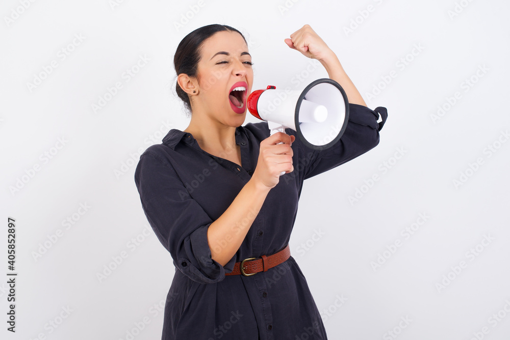 young beautiful Arab woman wearing gray dress against white studio background communicates shouting loud holding a megaphone, expressing success and positive concept, idea for marketing or sales. young beautiful Arab woman wearing gray dress against white studio background communicates shouting loud holding a megaphone, expressing success and positive concept, idea for marketing or sales.