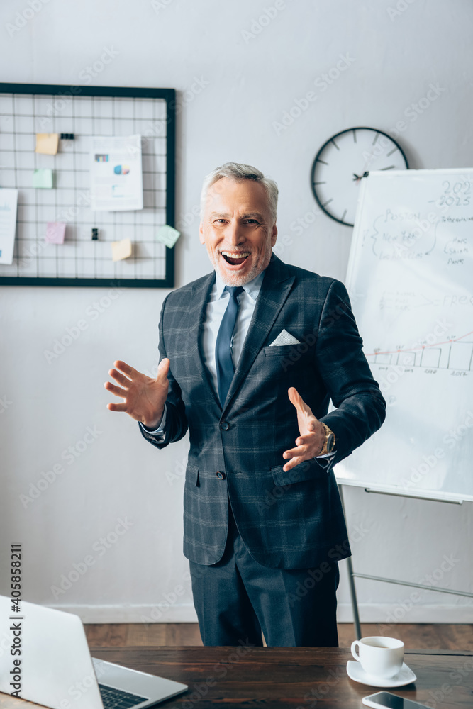Cheerful businessman in suit looking at camera near laptop and cup on blurred foreground