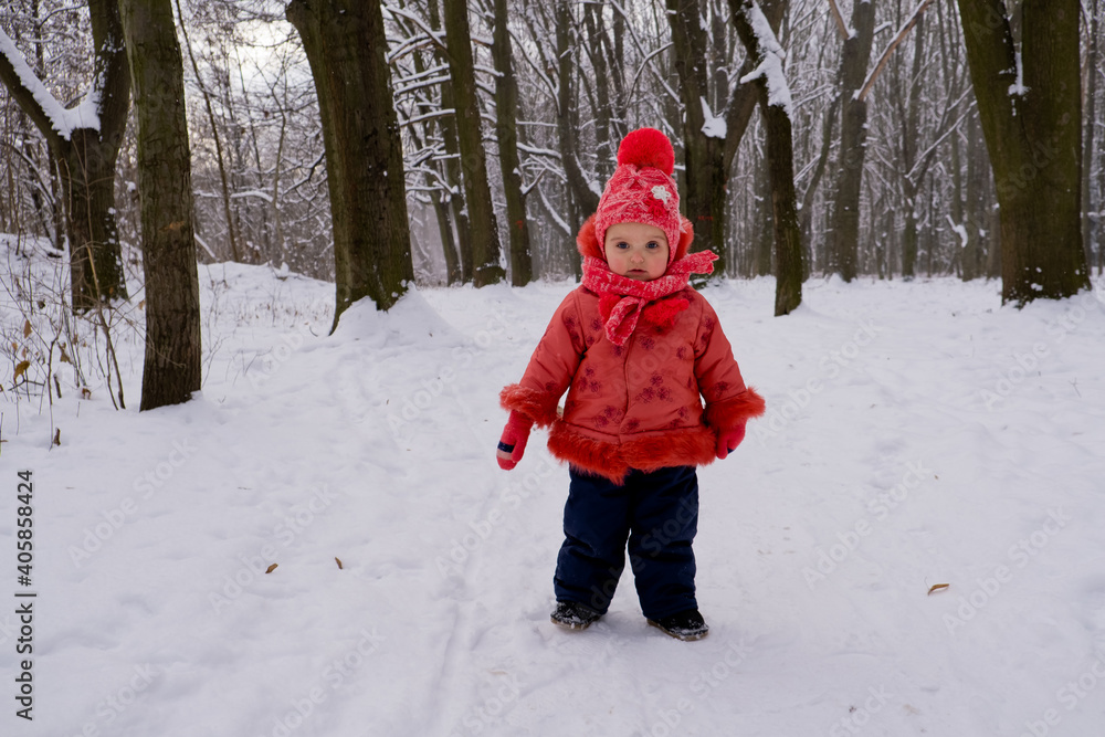 Baby girl in red jacket walking by snow in the forest.