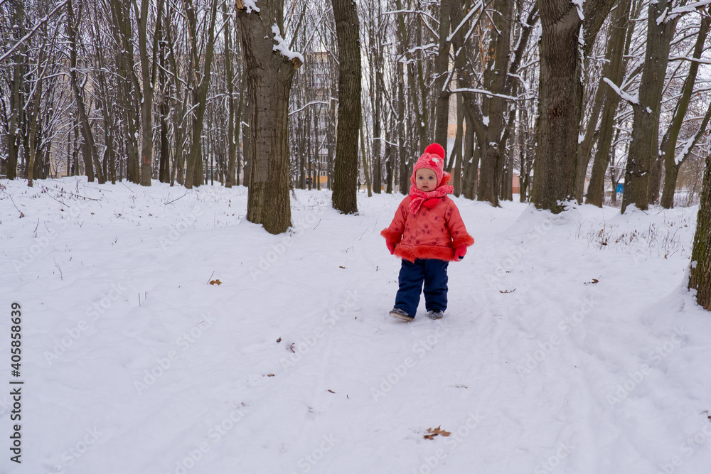 Baby girl in red jacket walking by snow in the forest.