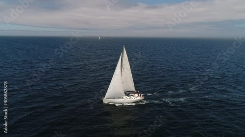 Aerial view. A white sailboat is moving fast on the waves of the sea. Flying around a boat in full sailing gear. Close hauled sailing in ocean. Yacht in windy and sunny day. Drone shot.