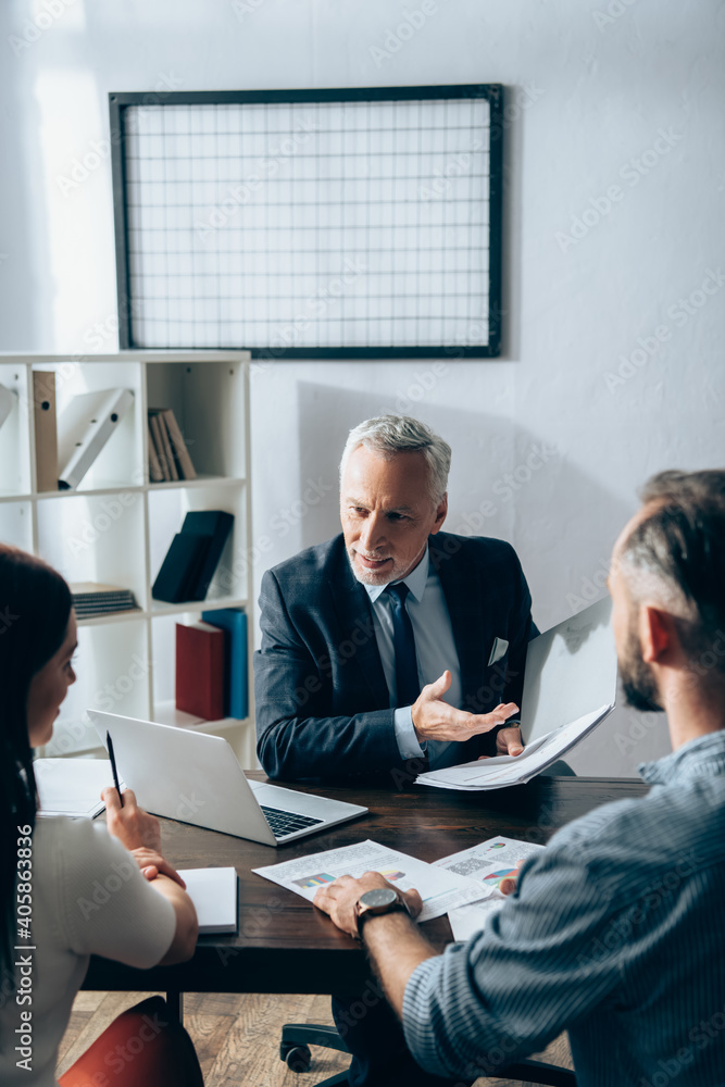 Obraz premium Smiling investor pointing with hand at paper folder near businesspeople on blurred foreground and laptop on table