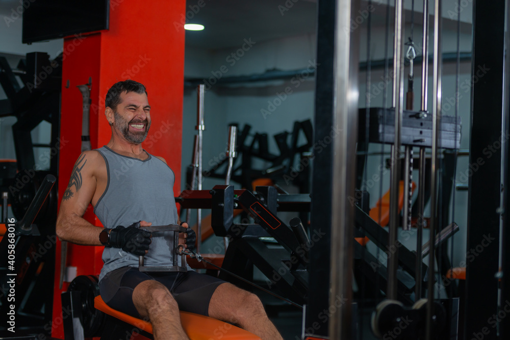 Fitness training in sport gym of active male with beard sitting and pulling heavy weight on exercise equipment Fitness training in sport gym of active male with beard sitting and pulling heavy weight on exercise equipment
