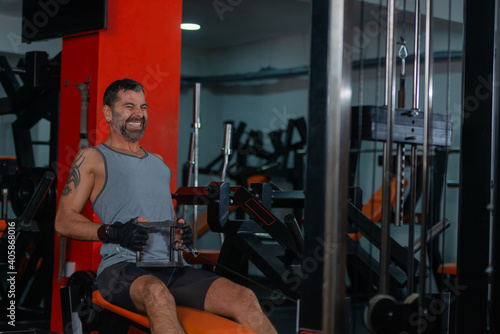 Fitness training in sport gym of active male with beard sitting and pulling heavy weight on exercise equipment