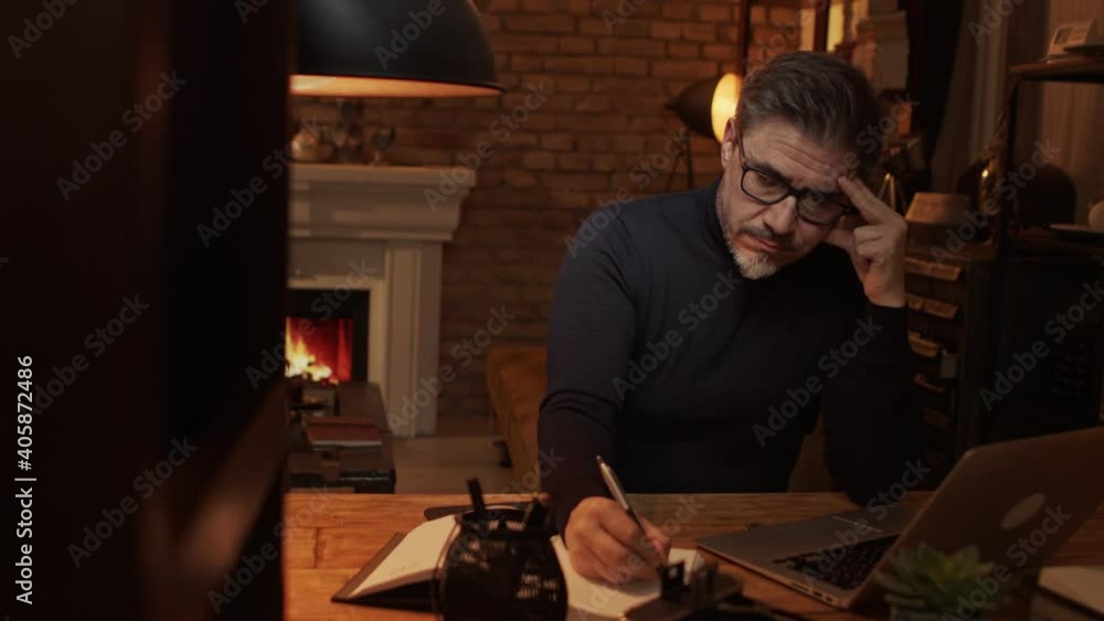 Home office - older white man working from home. Thinking, typing on laptop computer. Sitting at desk in dark living room iin front of fireplace. Home office - older white man working from home. Thinking, typing on laptop computer. Sitting at desk in dark living room iin front of fireplace.