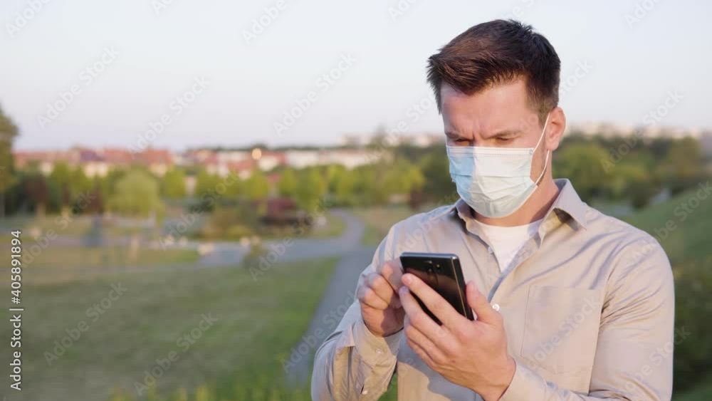 A young Caucasian man in a face mask works on a smartphone, then looks at the camera on a town's outskirts - buildings in the blurry background A young Caucasian man in a face mask works on a smartphone, then looks at the camera on a town's outskirts - buildings in the blurry background