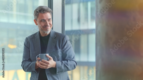 Happy older businessman taking break in office drinking coffee standing in window and daydreaming, thinking.