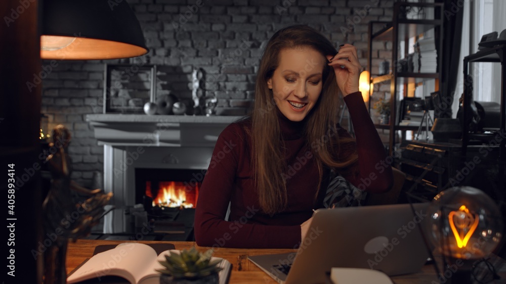 Home office - woman working from home with laptop computer. Talking on video chat, sitting at desk in dark living room in front of fireplace in Winter. Home office - woman working from home with laptop computer. Talking on video chat, sitting at desk in dark living room in front of fireplace in Winter.
