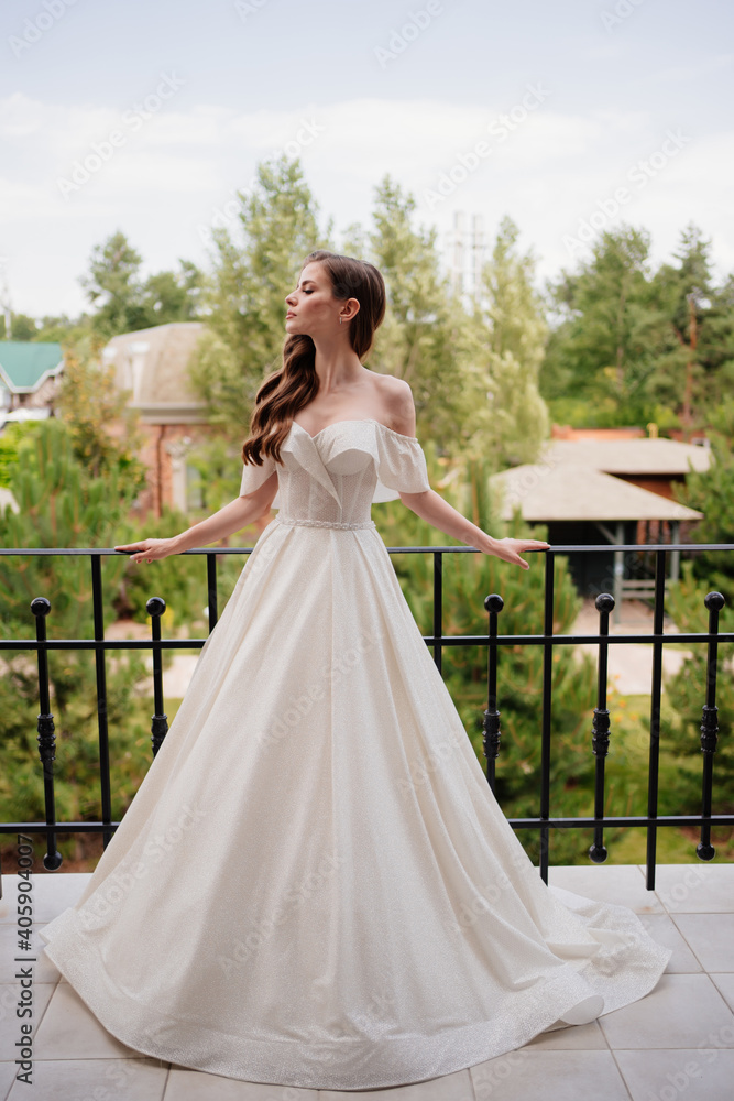 A beautiful young woman in a white wedding dress stands on the terrace. 