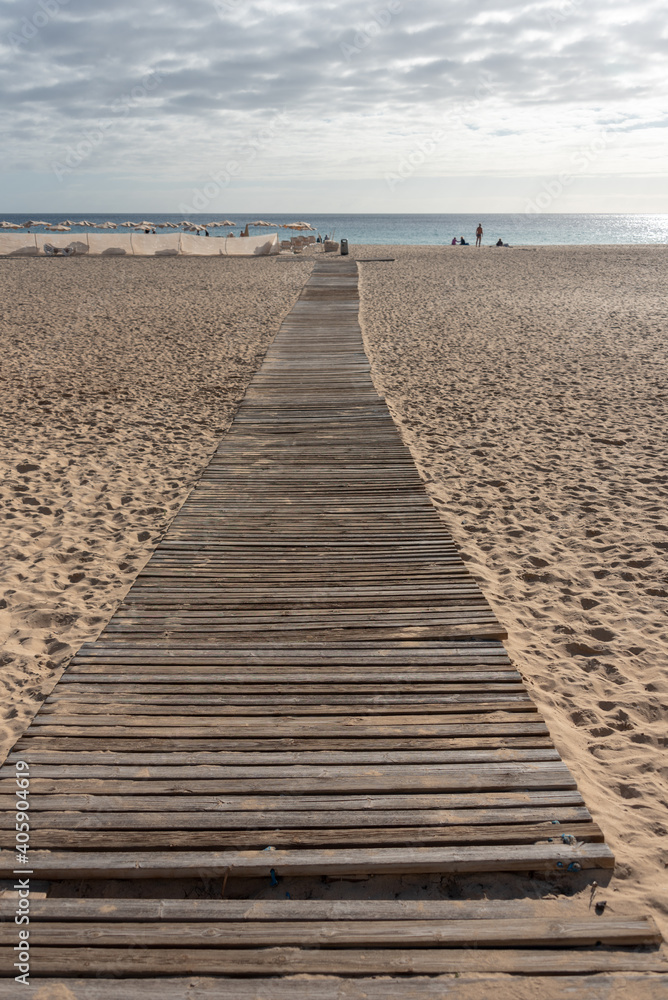 Wooden bridge on the sand in selective focus. Wide beach with umbrellas. Morro Jable, Jandia peninsula, Fuerteventura, Canary Islands. 