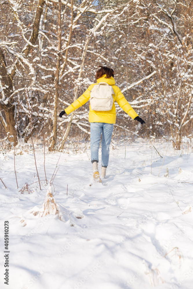 Back view of a woman in a bright yellow jacket and jeans with a backpack in a snowy landscape forest walks through the snowdrifts