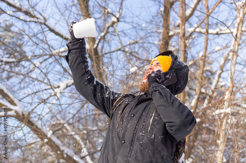 A brutal adult man in an orange bright hat in a snowy forest on a sunny day with a cup filled with snow, speaks by phone