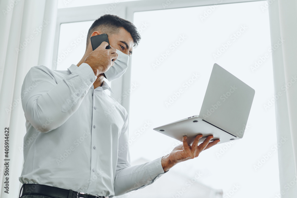 Handsome man businessman in medical mask using his laptop while talking on his cellphone Handsome man businessman in medical mask using his laptop while talking on his cellphone
