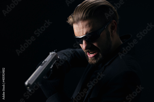 Aggressive bearded man in glasses, suit and gloves threatening someone with real gun. Isolated over black background. Concept of danger.