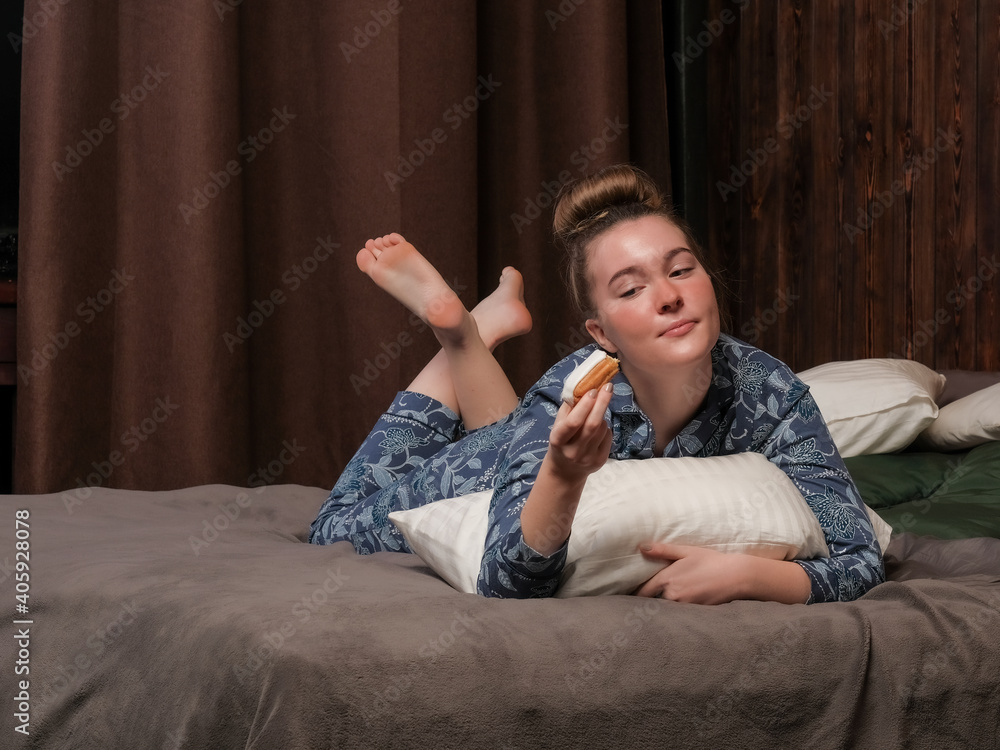 A young girl in pajamas lies on the bed and eats an eclair. Happy weekend at the hotel. Enjoyment of idleness.