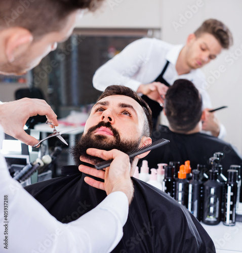 Satisfied male hairdresser accurately cutting beard of client at hair salon