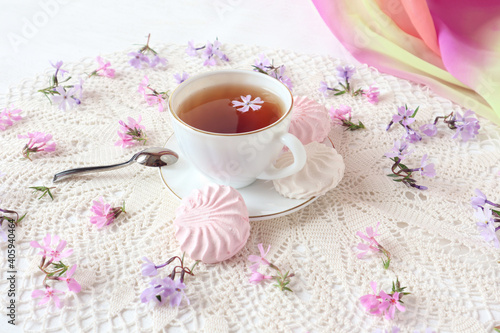 White and pink marshmallows on a plate with a cup of tea on a white openwork napkin, delicate flowers nearby, close-up
