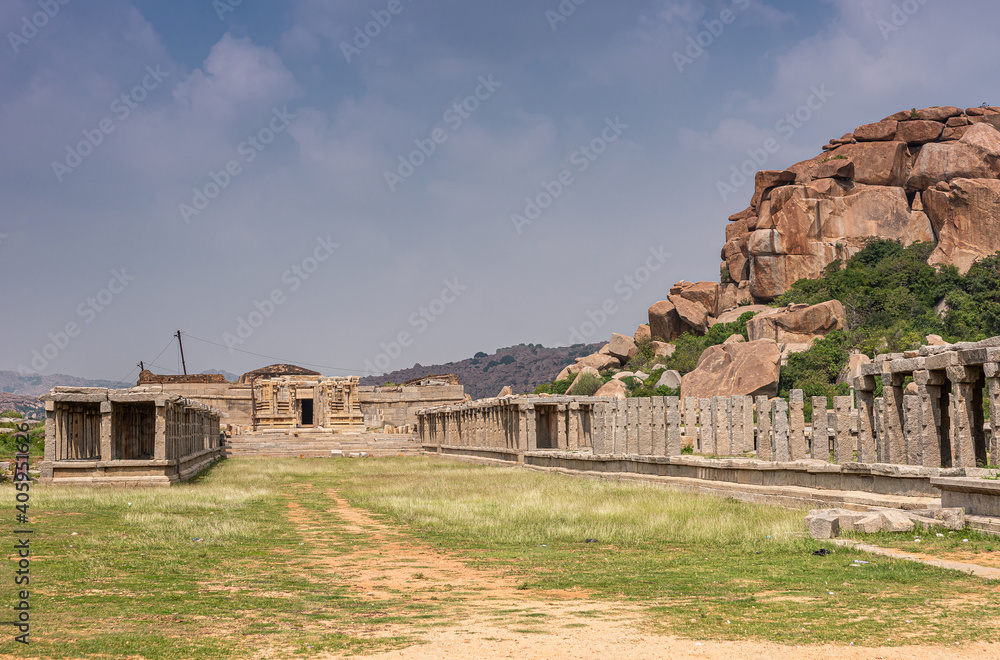 Hampi, Karnataka, India - November 5, 2013: Vijaya Vitthala Temple. Landscape outside shows green grass and ruinous galleries against brown boulder hill under blue cloudscape.