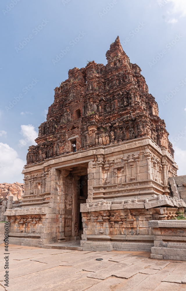Hampi, Karnataka, India - November 5, 2013: Vijaya Vitthala Temple. Red stone gopuram on top of beige stone east entrance gate under blue cloudscape.