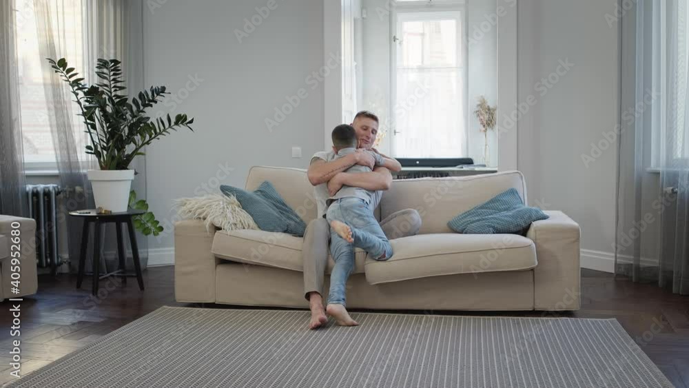 Young Man Sitting On Sofa In Apartment. His Son Runs To Him. Young Man Sitting On Sofa In Apartment. His Son Runs To Him.