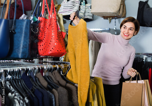 Happy cheerful positive female shopper examining warm sweaters in women cloths shop