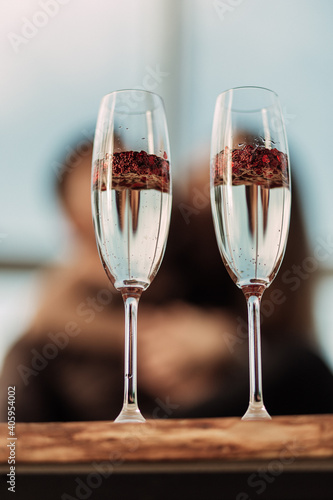 Happy Valentines day celebration concept. Close up of 2 two glasses with sparkling champagne and red heart shaped confetti. Love potion in glass. Loving couple blurred on background