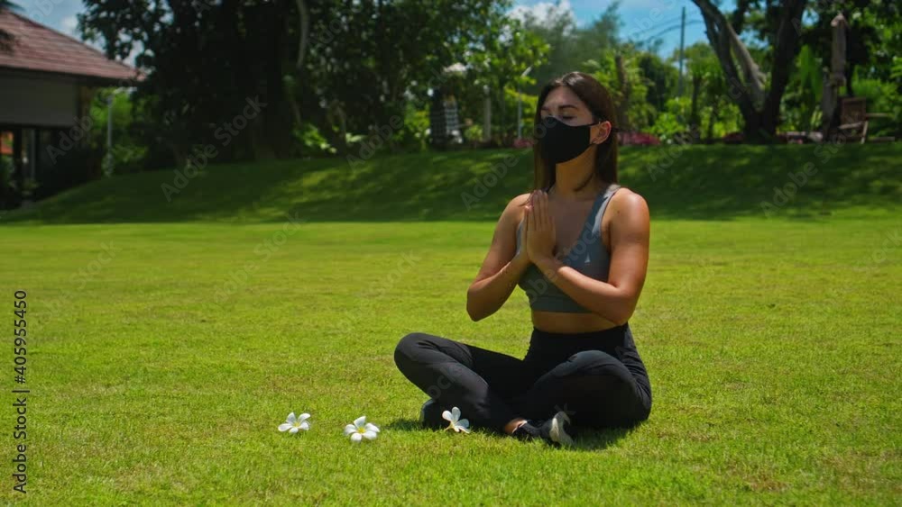 Young European brunette girl practices alone yoga in nature sitting on green grass in lotus pose wearing a black protective mask Young European brunette girl practices alone yoga in nature sitting on green grass in lotus pose wearing a black protective mask