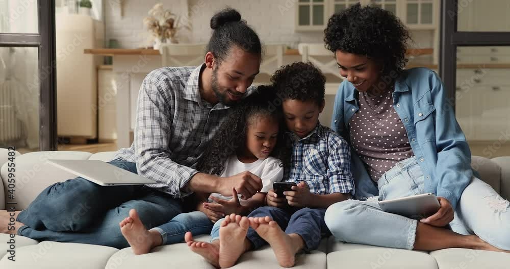 Young African family with siblings kids sit on couch with diverse gadgets, hold tablet laptop and cellphone. Usage of mobile application, bad habits, spend free time use modern tech, overuse concept Young African family with siblings kids sit on couch with diverse gadgets, hold tablet laptop and cellphone. Usage of mobile application, bad habits, spend free time use modern tech, overuse concept