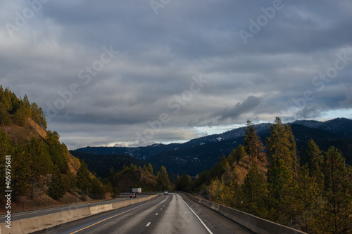 Highway among tall fir trees and blue mountains in front, against a gray sky in the clouds