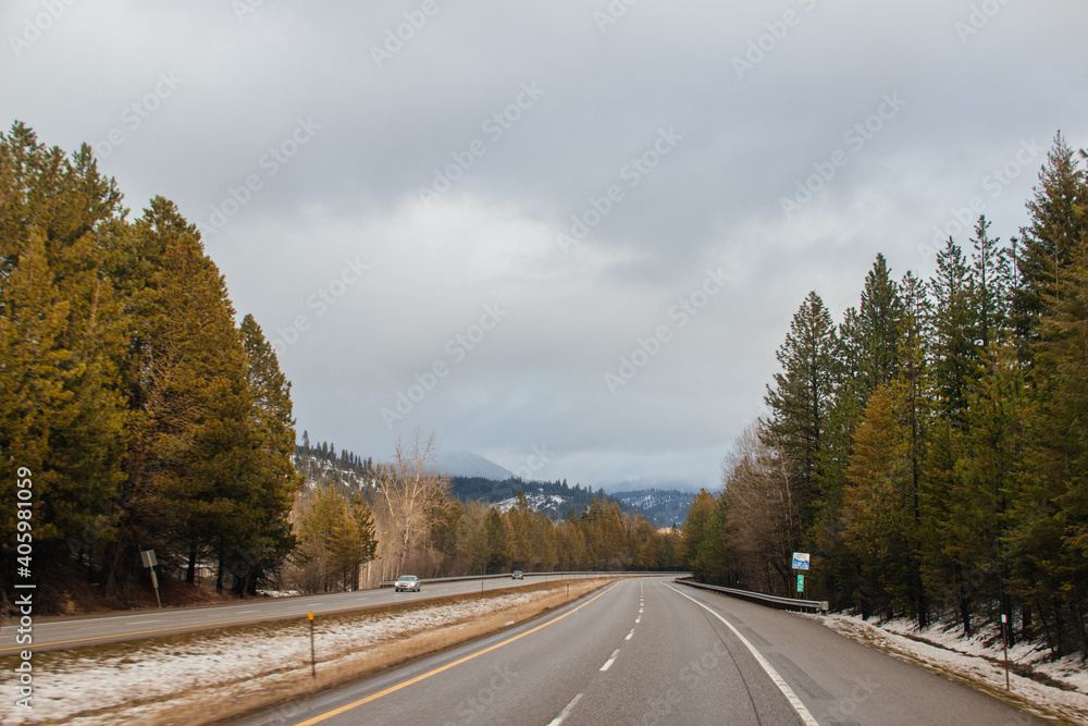 Winter landscape -a highway along which cars drive, surrounded by tall trees, on which the rays of the setting sun fall, blue mountains covered with snow are visible in front.Kingston,ID,USA,2-8-2020