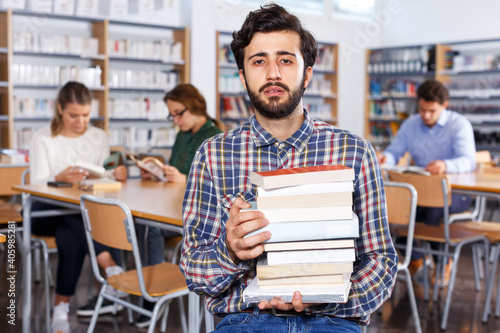 Sad tired man engaged in research sitting in busy library with pile of books
