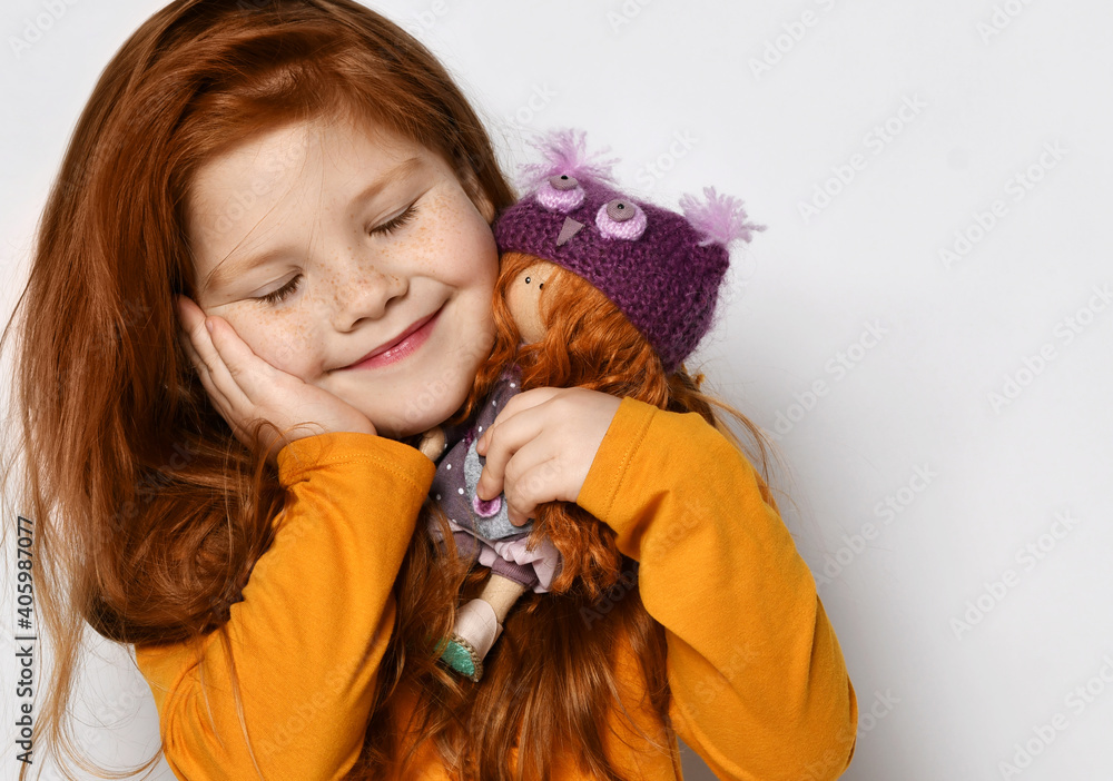 Portrait of sleeping and smiling five-year-old red-haired kid girl with closed eyes in orange sweatshirt holding small redhair doll in hands at her face over white background with copy space Portrait of sleeping and smiling five-year-old red-haired kid girl with closed eyes in orange sweatshirt holding small redhair doll in hands at her face over white background with copy space
