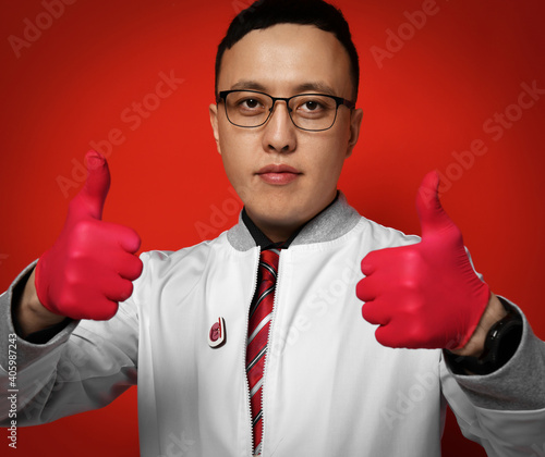 Portrait of young man doctor urologist or proctologist in medical gown and red latex gloves gesturing thumbs up good condition sign after treatment over red background. Urology proctology concept photo